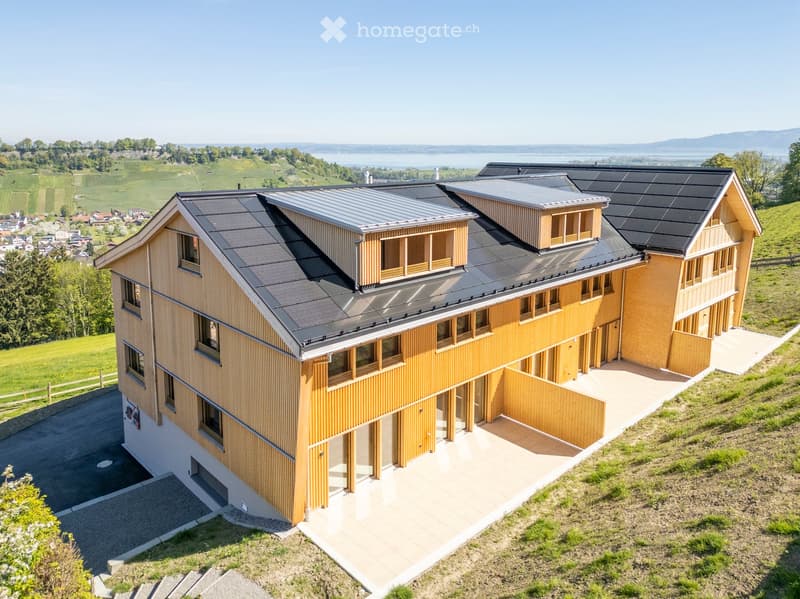 Modern two-story house with wooden siding, grey metal roof, large windows, and solar panels on the roof. The house is situated on a hillside with a scenic view of a lake and surrounding landscape.