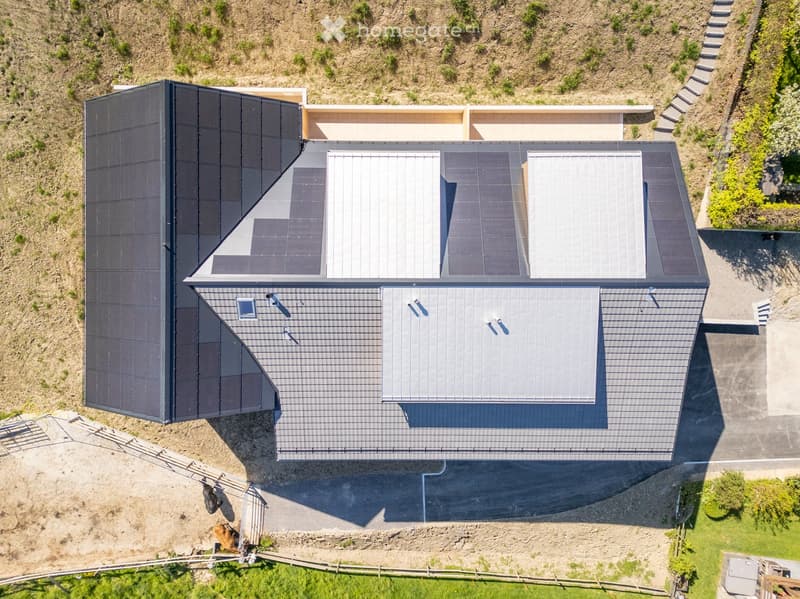 Aerial view of a modern house with a grey metal roof, solar panels, and a surrounding landscape with grass and trees.