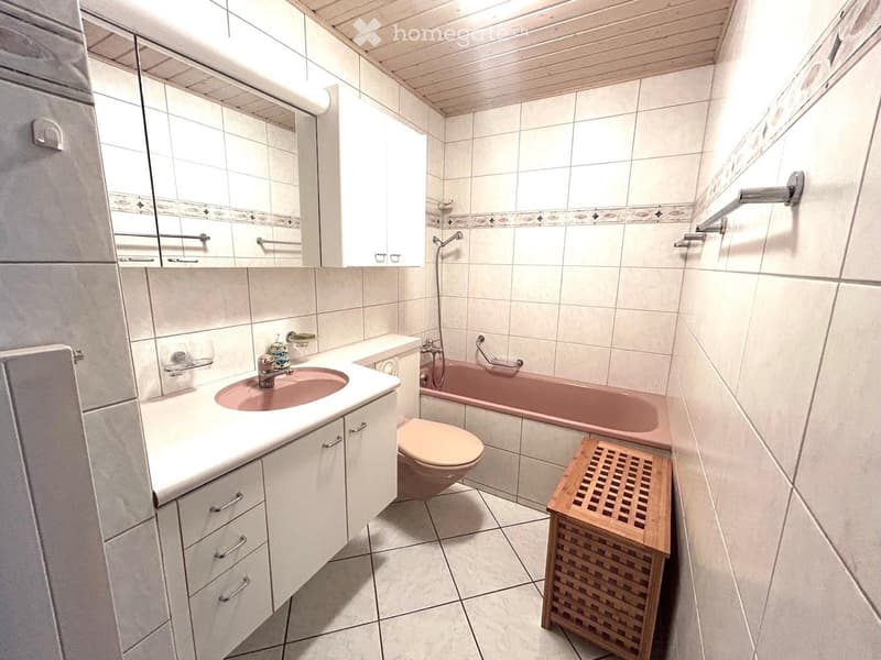 Bathroom with white tiled walls and floor, featuring a sink with a pink basin, a toilet, a pink bathtub, a wooden stool, and a mirrored cabinet.