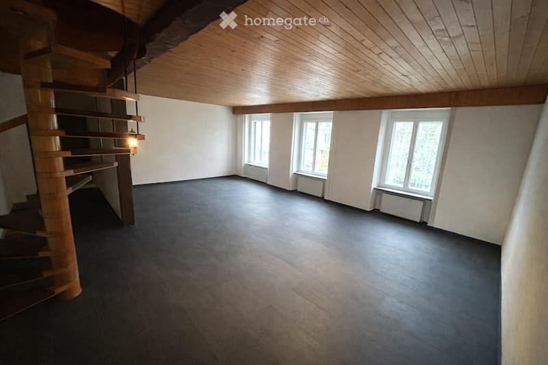 Empty room with wooden ceiling, dark tiled floor, white walls, and a wooden staircase leading upwards. Three windows with white frames and radiators beneath them.