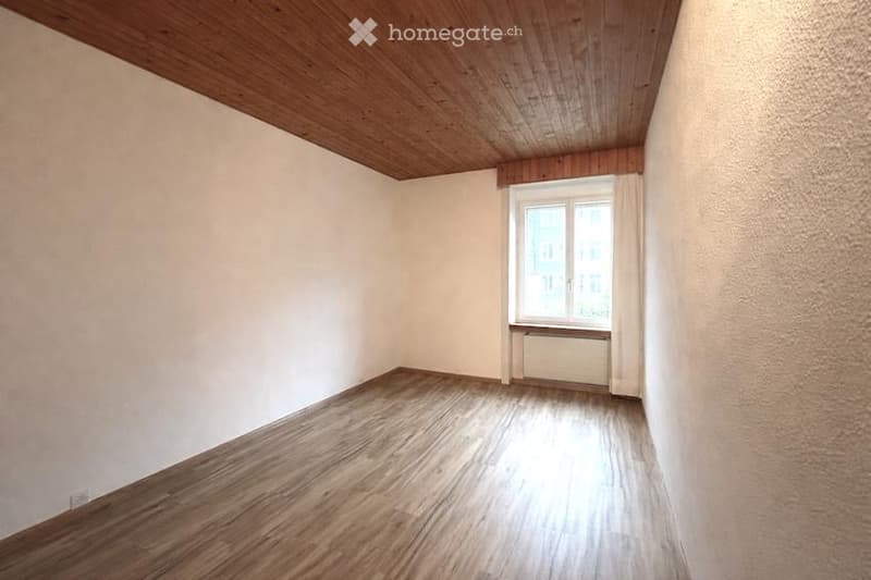 Empty room with wooden flooring, white walls, and a single window with white curtains. The ceiling features wooden paneling.