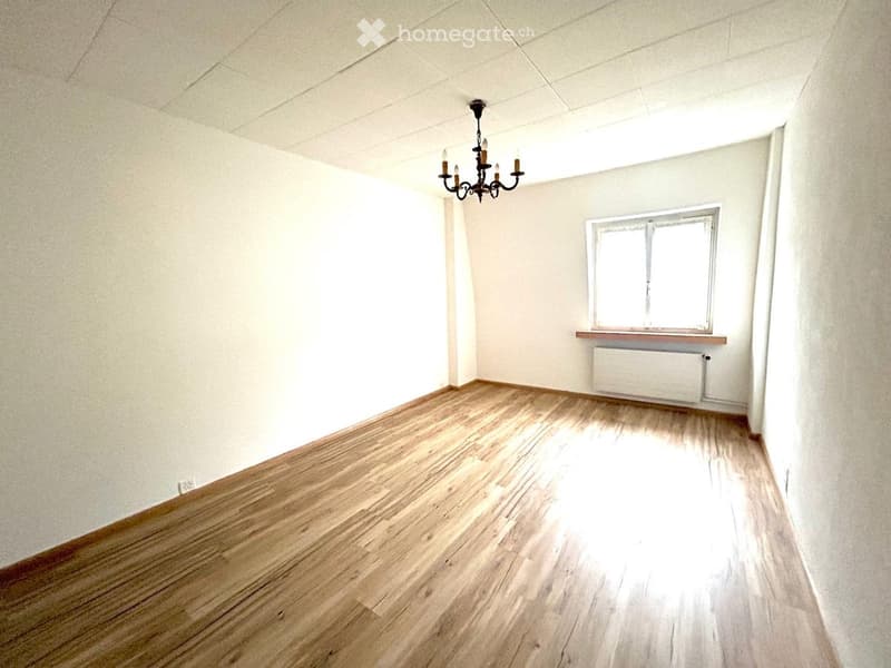 Empty room with wooden flooring, white walls, a window with a radiator below, and a chandelier hanging from the ceiling.
