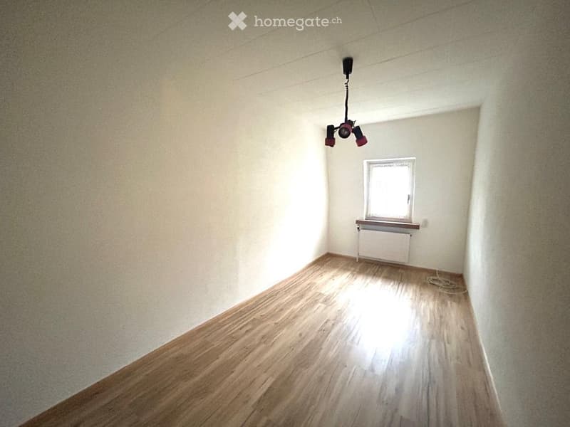 Empty room with wooden flooring, white walls, a single window with a radiator below, and a ceiling light fixture.