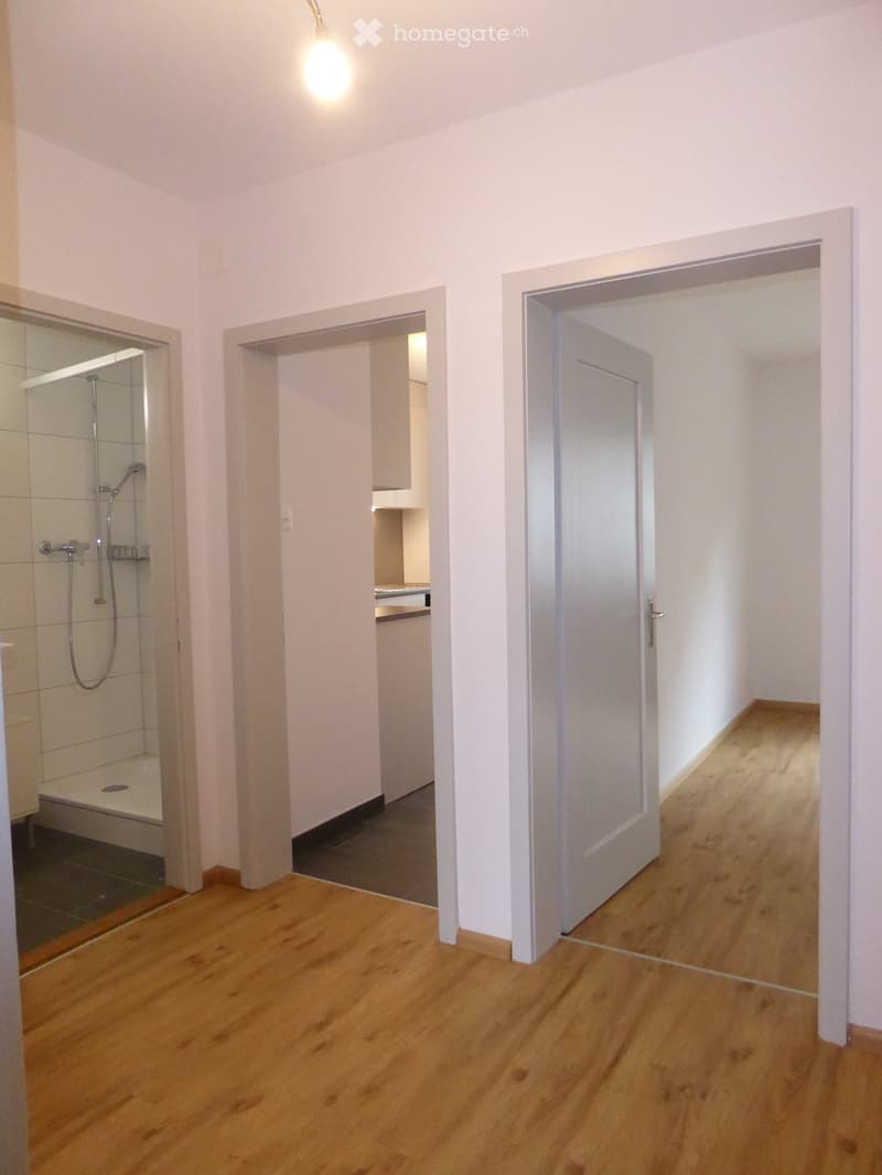 Empty hallway with wooden flooring, white walls, and open doors leading to a bathroom and another room.