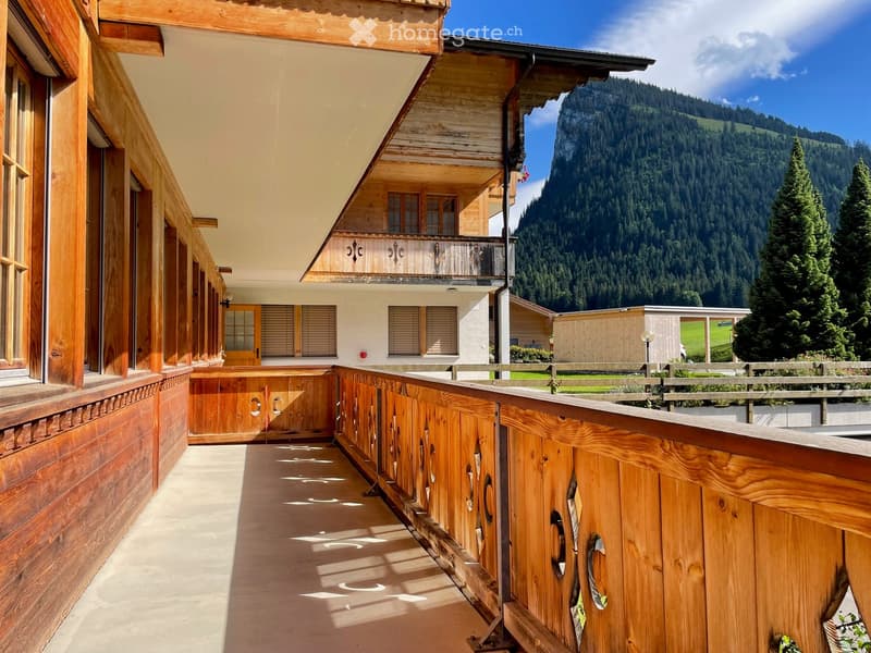 Wooden balcony with a wooden railing, overlooking a scenic mountain landscape with trees and a clear blue sky.