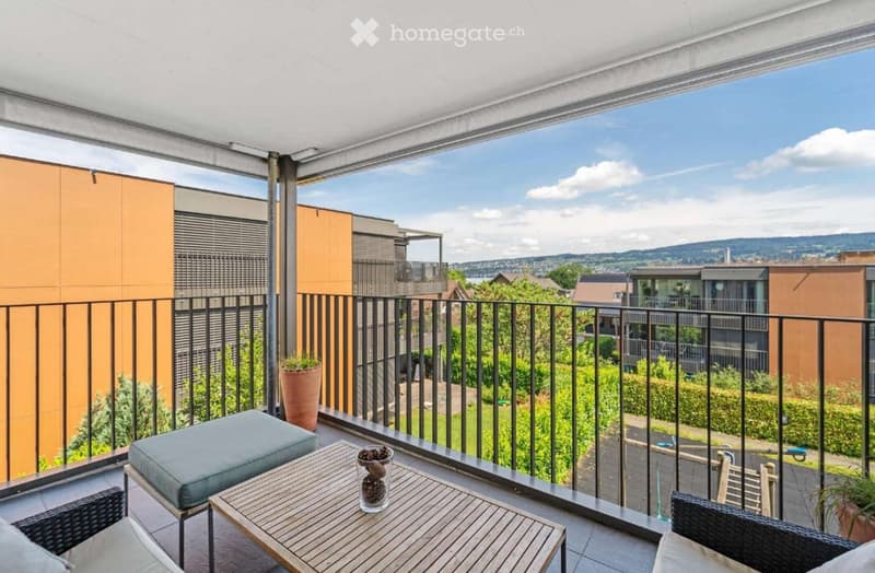 Balcony with wooden table, grey cushioned stool, wicker chairs, and potted plants, overlooking a green area with modern buildings.