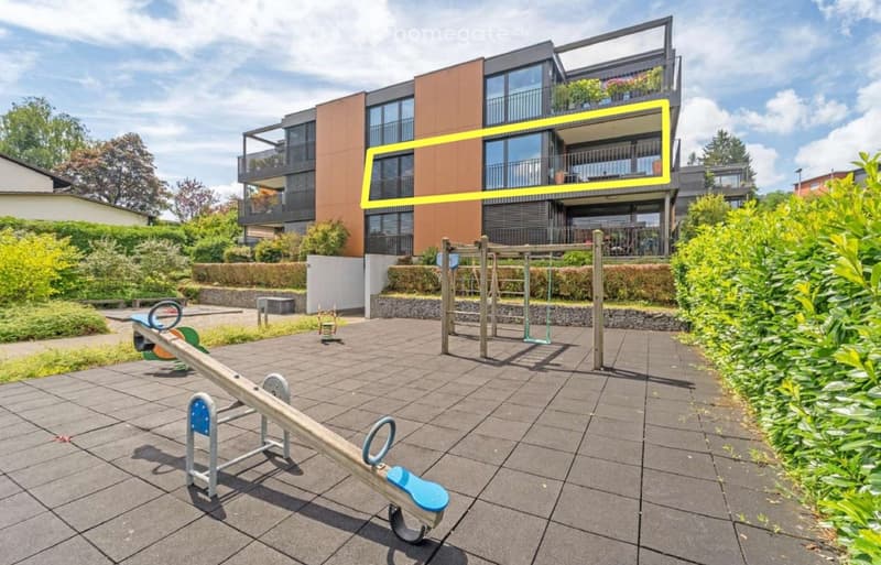 Outdoor playground area with a seesaw, climbing frame, and surrounding greenery, adjacent to a modern multi-story residential building with balconies.