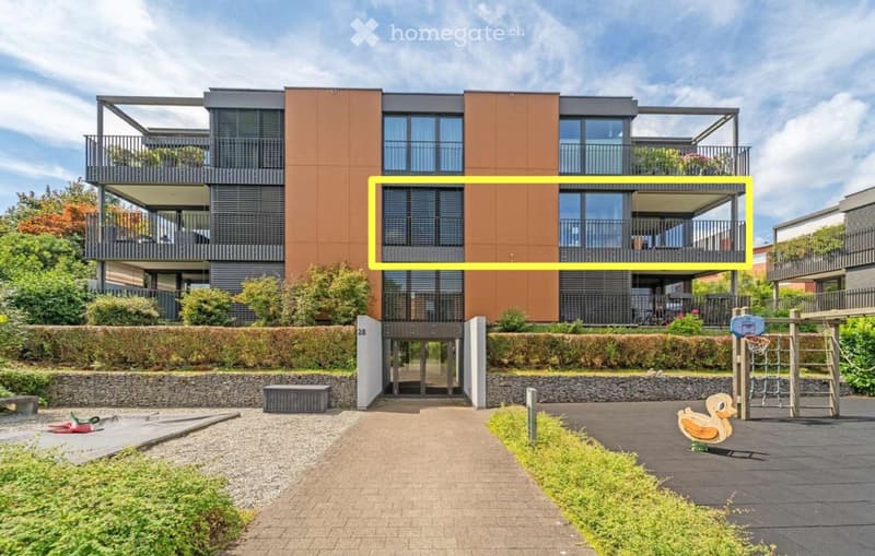 Modern multi-story residential building with balconies, large windows, and a landscaped entrance area featuring a playground and greenery.