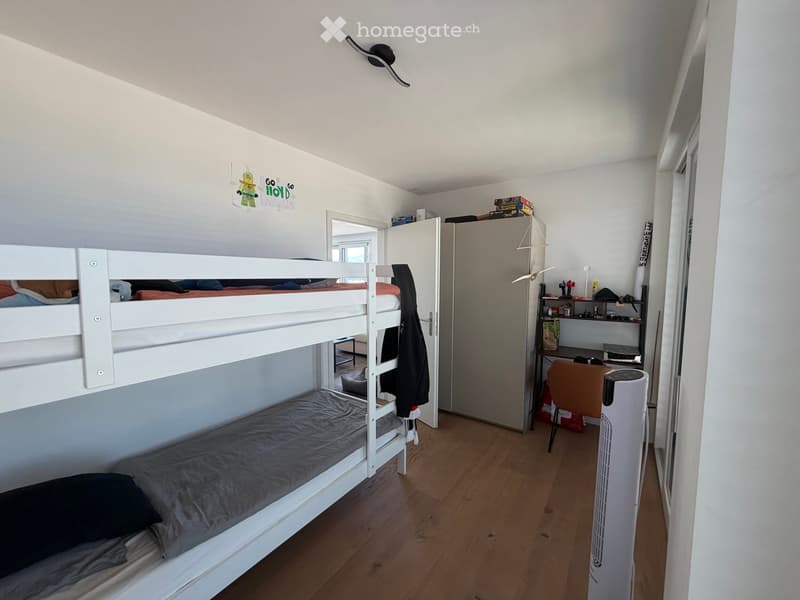 A furnished bedroom with a bunk bed, a desk with a chair, a fan, and a cabinet. The room has wooden flooring and white walls.
