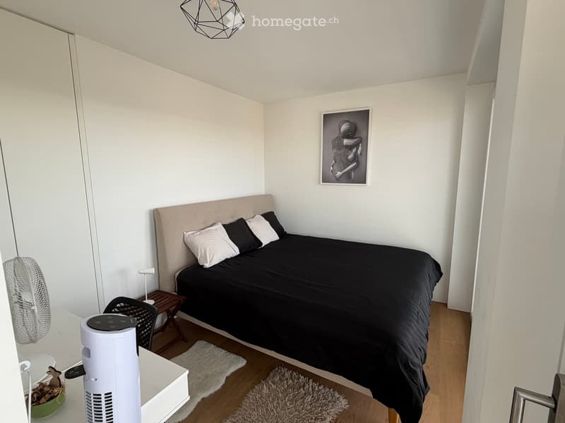 A modern bedroom with a large bed, nightstand, fan, chair, and a picture frame on the wall.