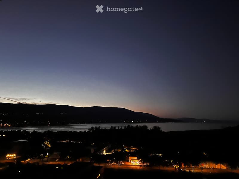 Nighttime view of a town with a lake and mountains in the background, illuminated by streetlights and building lights.