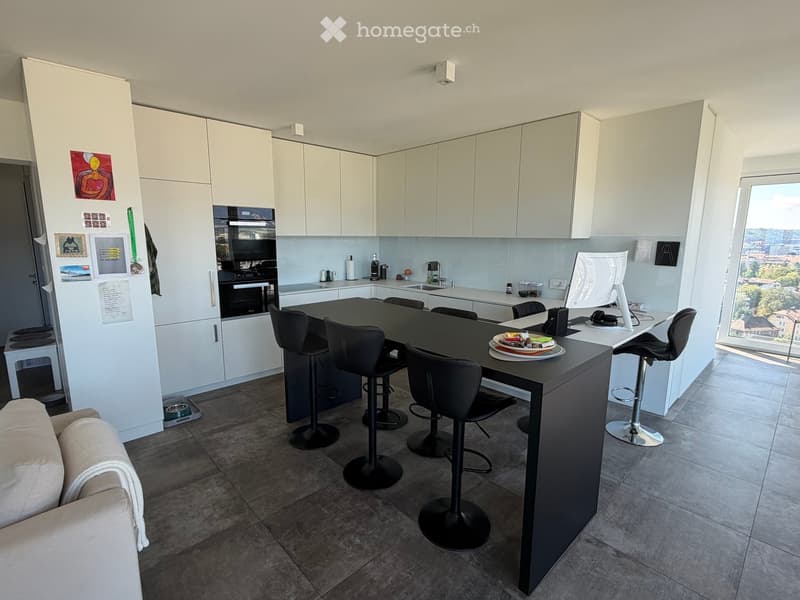Modern kitchen with white cabinets, a central island, bar stools, and a sofa in the foreground. The kitchen features a refrigerator, oven, sink, and a laptop on the island.