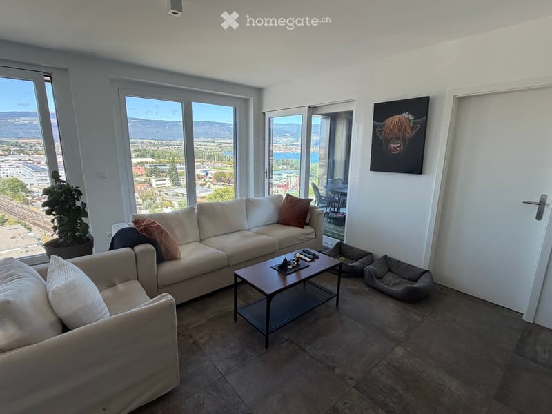 Modern living room with large windows offering a scenic view, featuring a beige sectional sofa, a wooden coffee table, a potted plant, and a picture frame on the wall.