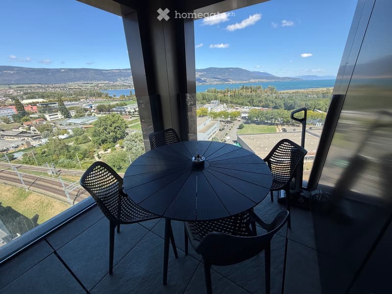Balcony with a round wooden table and four black mesh chairs, offering a panoramic view of a cityscape, water body, and distant mountains.
