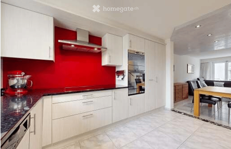 Modern kitchen with white cabinets, red accent wall, and stainless steel appliances. Includes a mixer on the counter and a dining area with a table and chairs.