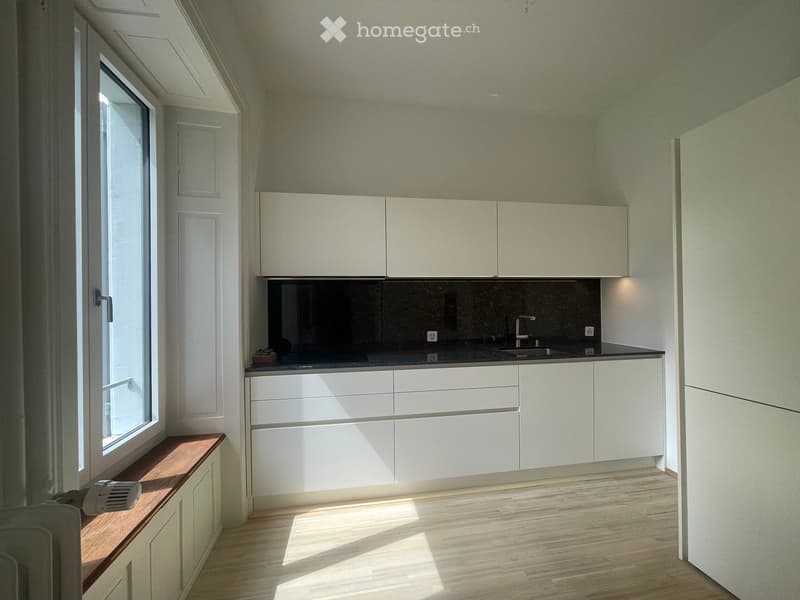 Modern kitchen with white cabinets, dark countertop, and a window bench. The space is bright with natural light and has a sleek, minimalist design.