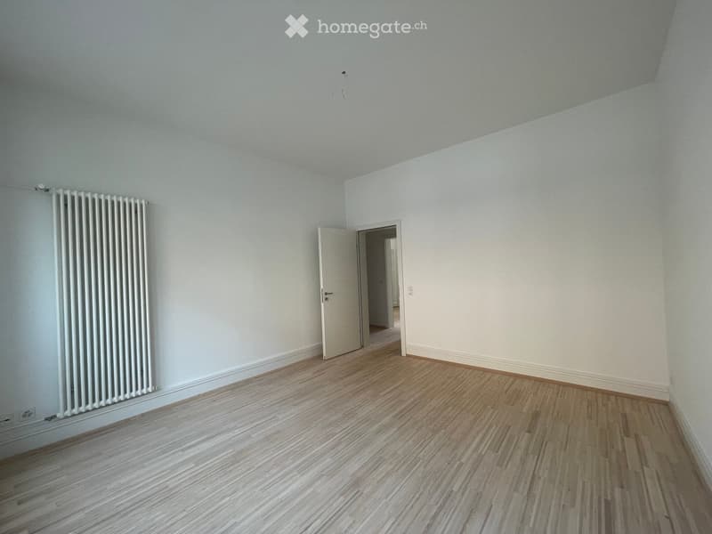 Empty room with light blue walls, wooden flooring, and a white radiator on one wall. An open door leads to another room.