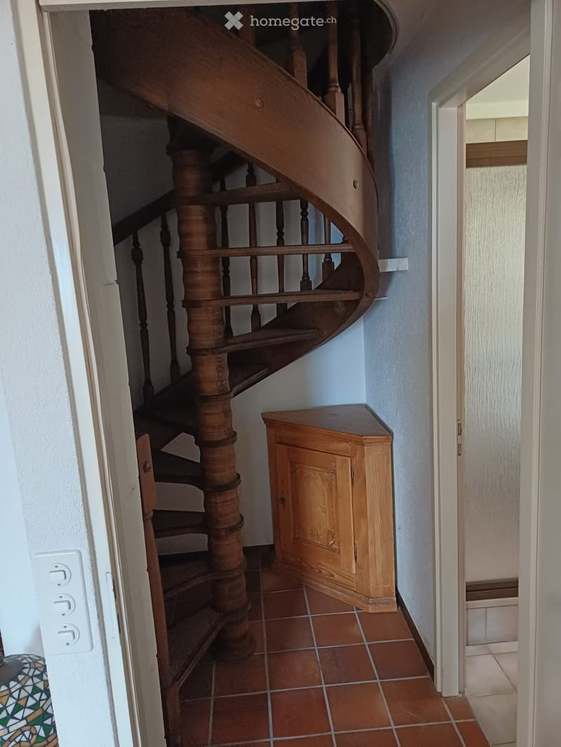 A narrow hallway with a wooden spiral staircase, light blue walls, and a small wooden cabinet on the right side.