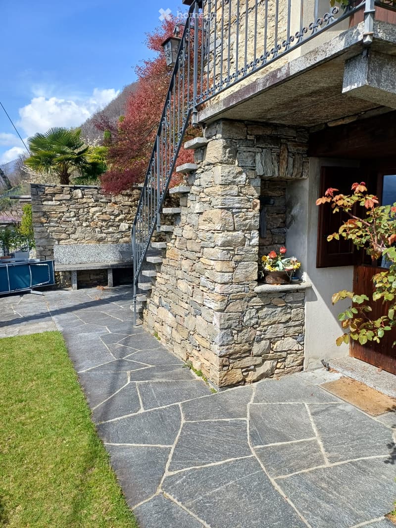 Stone pathway leading to a house entrance with a stone wall, metal railing, and small garden area with potted plants.