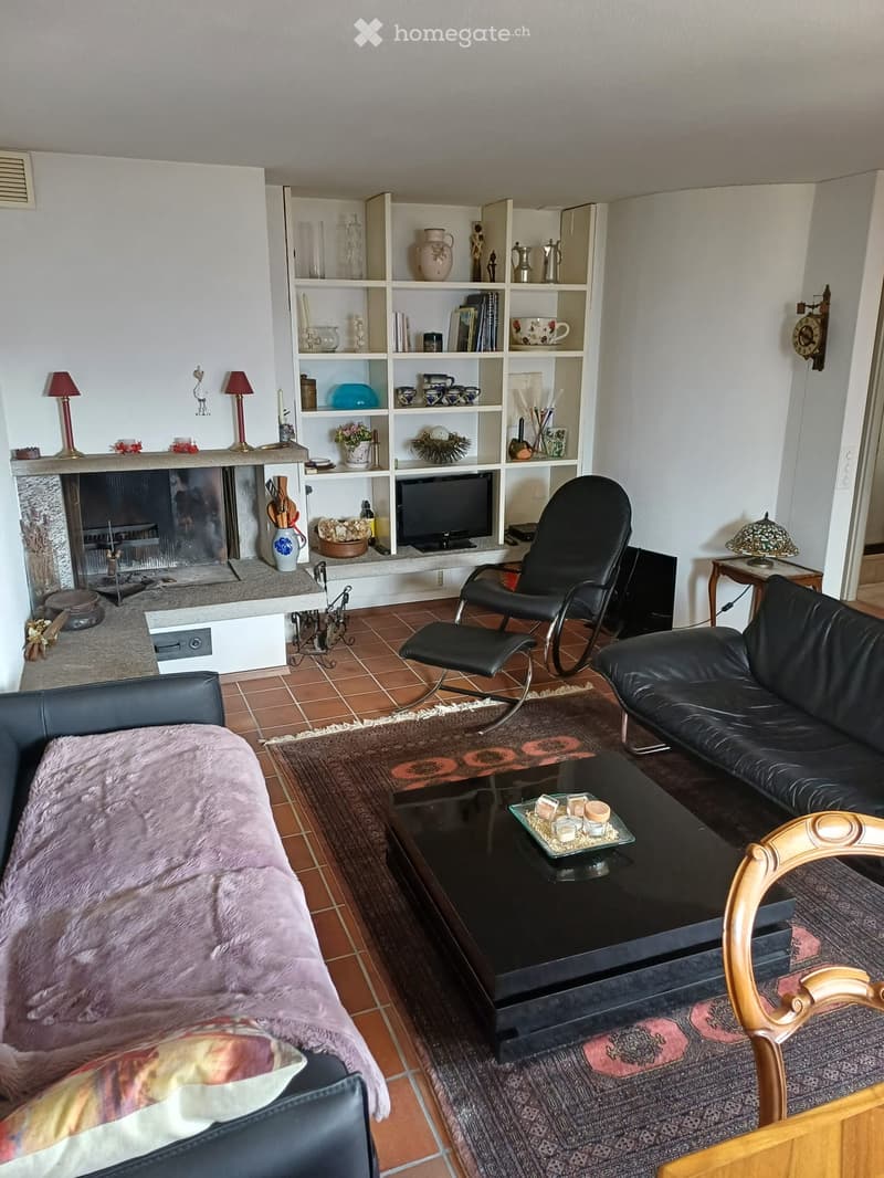 Living room with a fireplace, tiled floor, and various seating arrangements including a sofa, chair, and ottoman. Shelves hold decorative items and books, and a coffee table sits in the center.