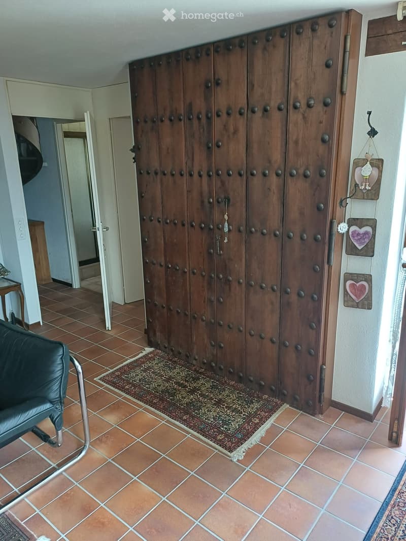 Hallway with terracotta tiles, large wooden door with decorative knobs, small table, chair, and wall decorations.