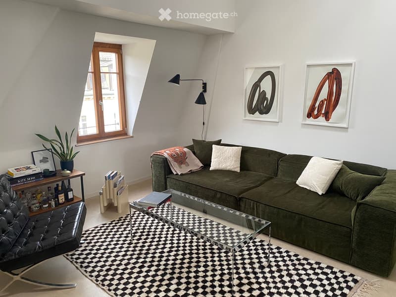 Modern living room with a green sectional sofa, glass coffee table, black leather chair, side table with books and bottles, and artwork on the walls.