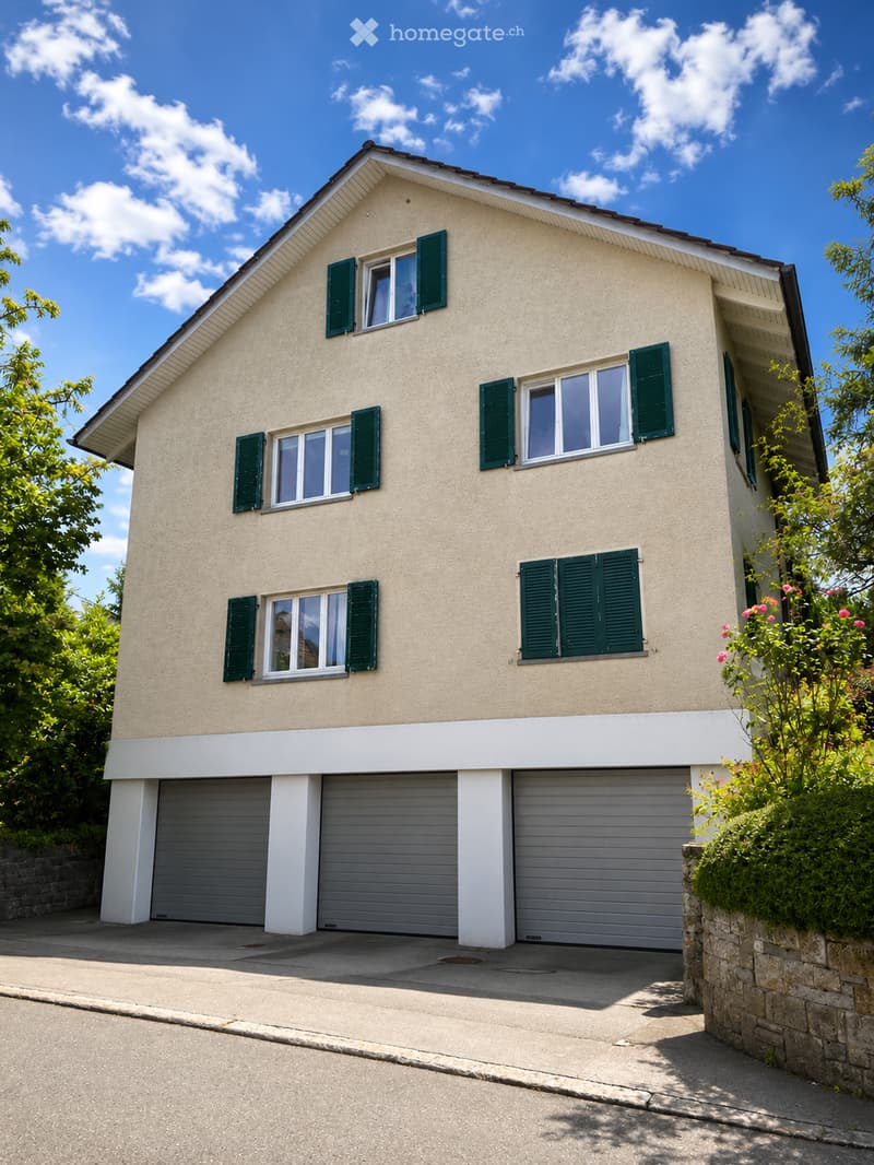 Two-story house with beige siding, green shutters, multiple windows, and a three-car garage.