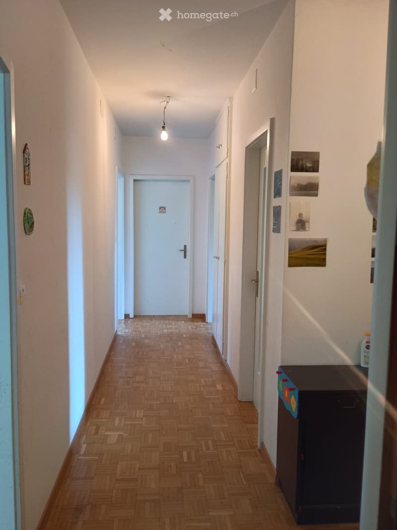 A narrow hallway with wooden flooring, white walls, and several doors. There are framed pictures on the right wall and a small table with items on it.