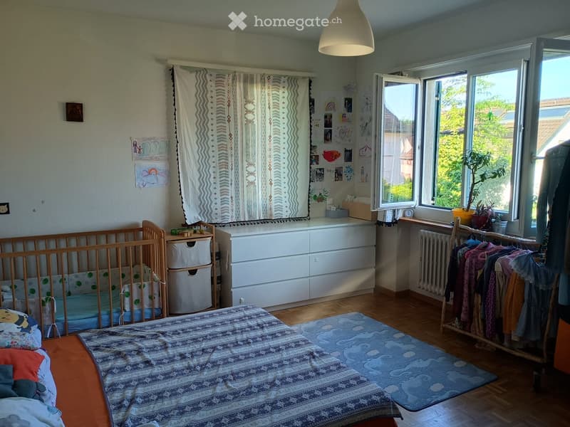 Brightly lit bedroom with a large bed, a crib, a wardrobe, and a window with curtains. The room features wooden flooring, a radiator, and several decorative items including drawings and a plant on the windowsill.