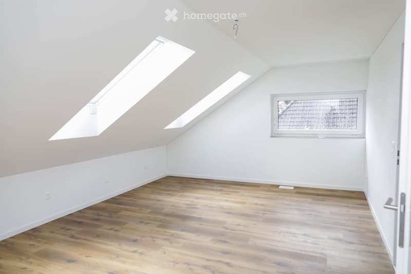 Empty attic room with wooden flooring, white walls, skylights, and a window showing a roof view.