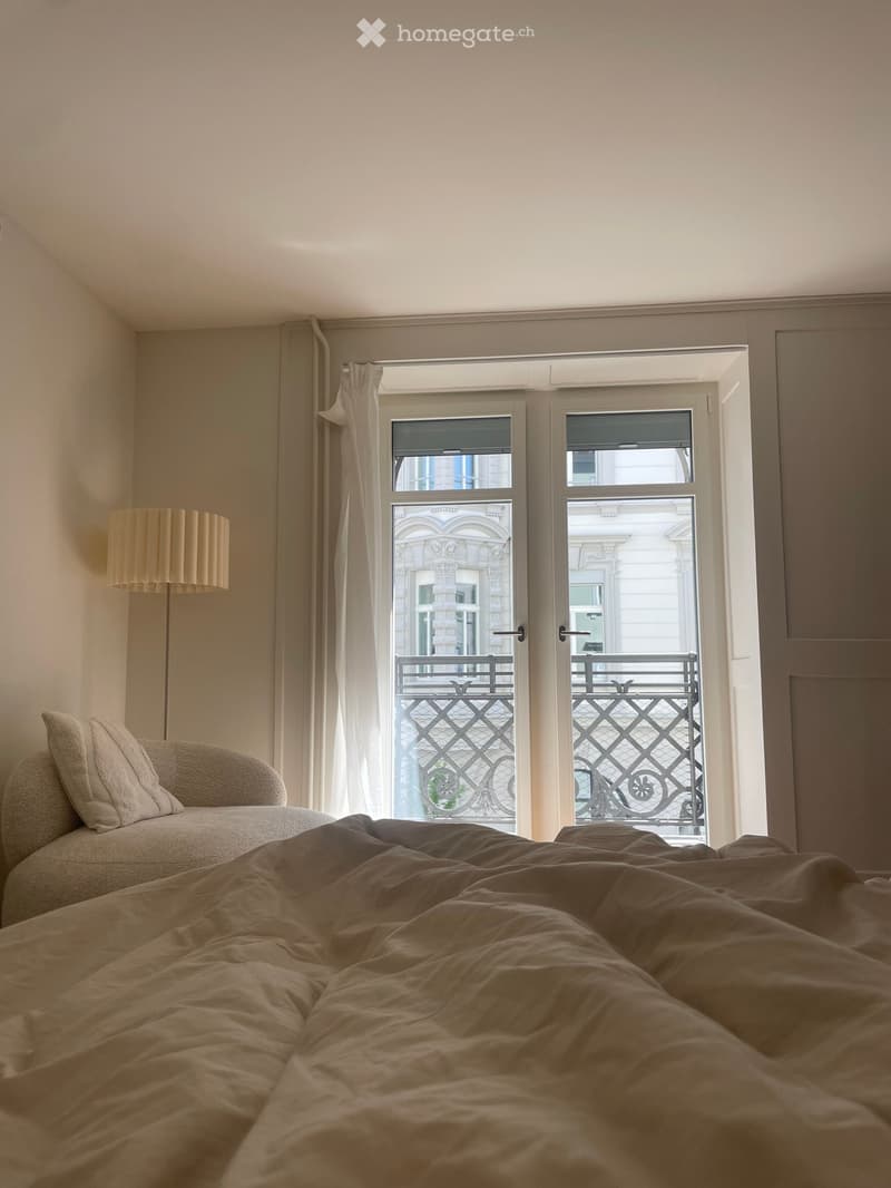 Cozy bedroom with a large bed, sofa, and floor lamp near a large window with a view of a balcony.
