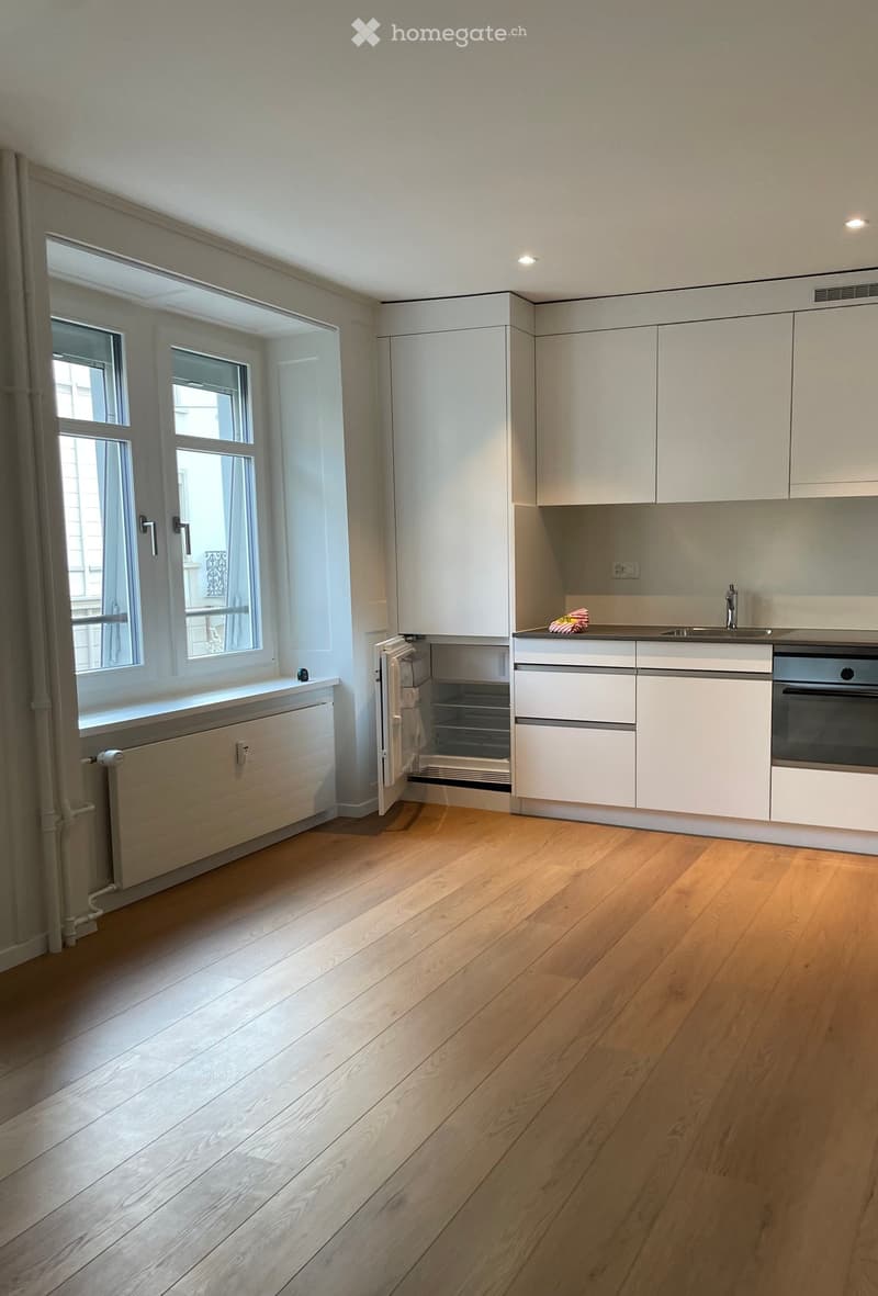 Modern kitchen with light-colored cabinets, wooden flooring, and large windows allowing natural light. Features include a sink, faucet, oven, and refrigerator.