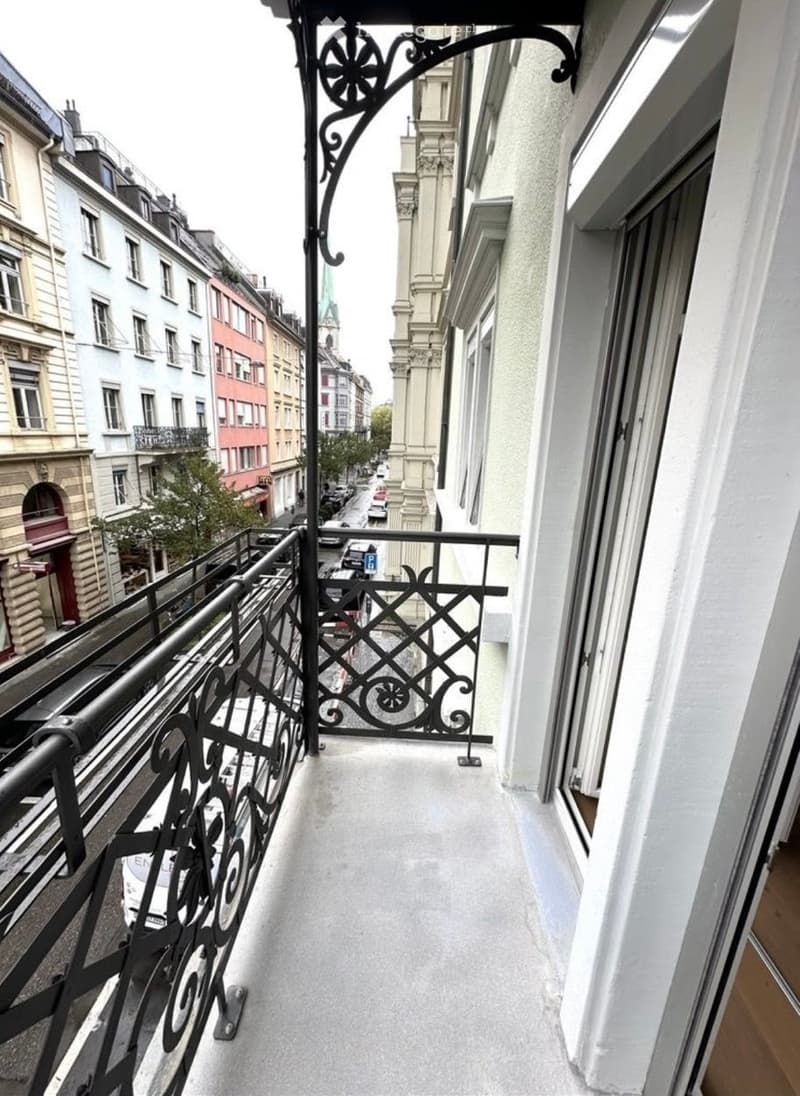 Balcony with ornate black metal railing, overlooking a city street with buildings and parked cars.