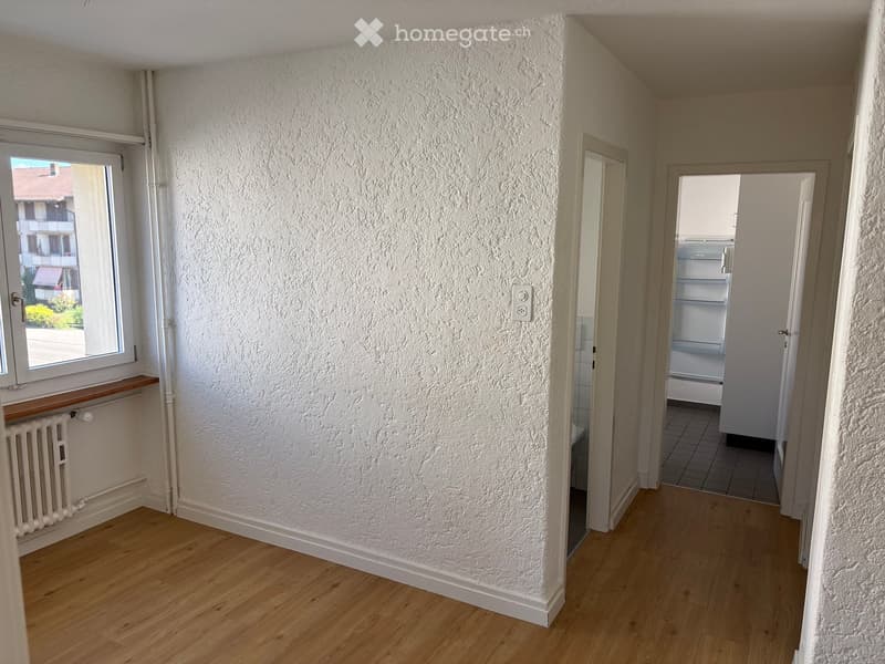 Empty hallway with wooden flooring, white textured walls, a window with a view of a building, and a radiator under the window. A doorway leads to another room with a visible refrigerator.
