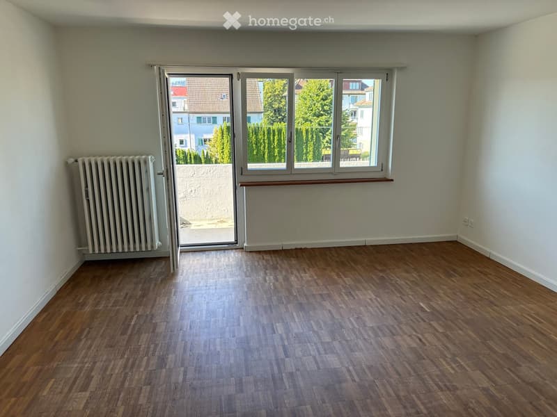 Empty room with wooden flooring, white walls, a radiator, and large windows with a view of trees and buildings outside.