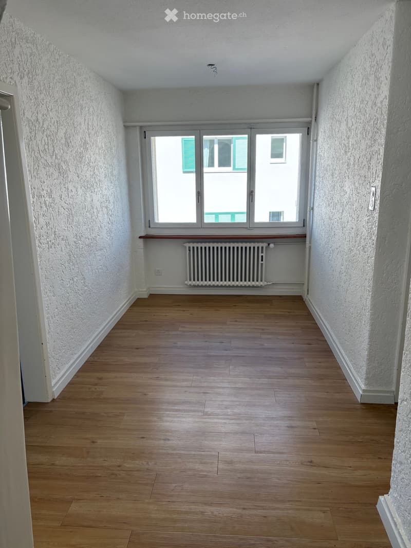Empty hallway with wooden flooring, white walls, and a window with shutters. A radiator is placed below the window.