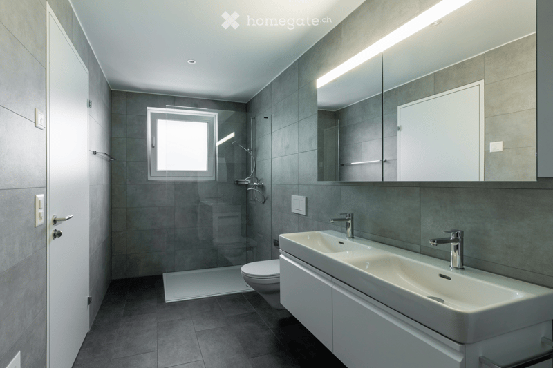 Modern bathroom with grey tiled walls and floor, double sink with chrome faucets, large mirror, and a glass-enclosed shower area.