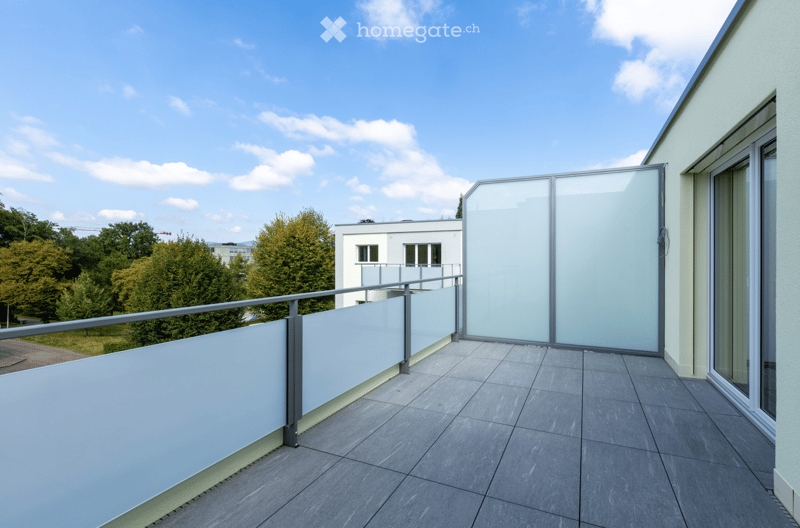 Modern balcony with grey tiled floor, glass balustrade, and sliding glass panels, overlooking a residential area with trees and buildings.