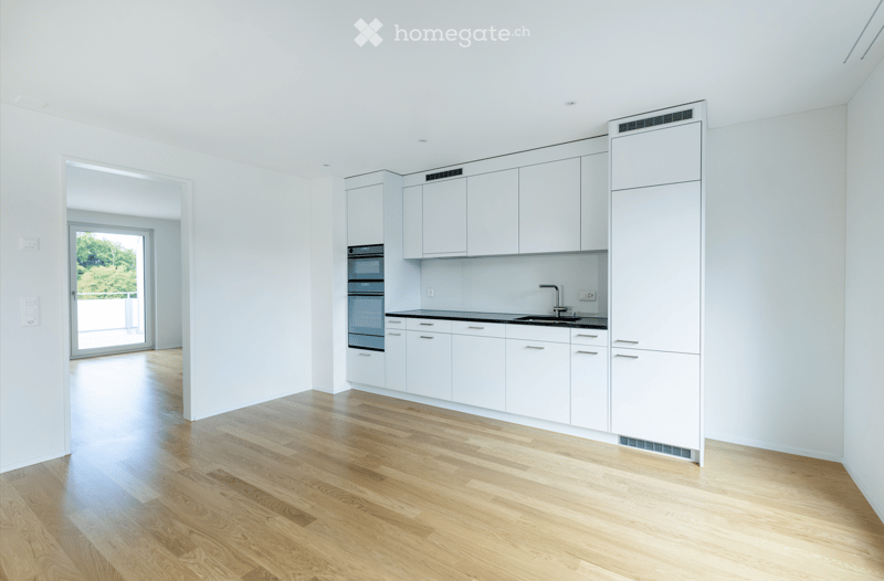 Modern, unfurnished kitchen with white cabinets, stainless steel appliances, and a wooden floor. The space is bright with large windows and a door leading to an outdoor area.