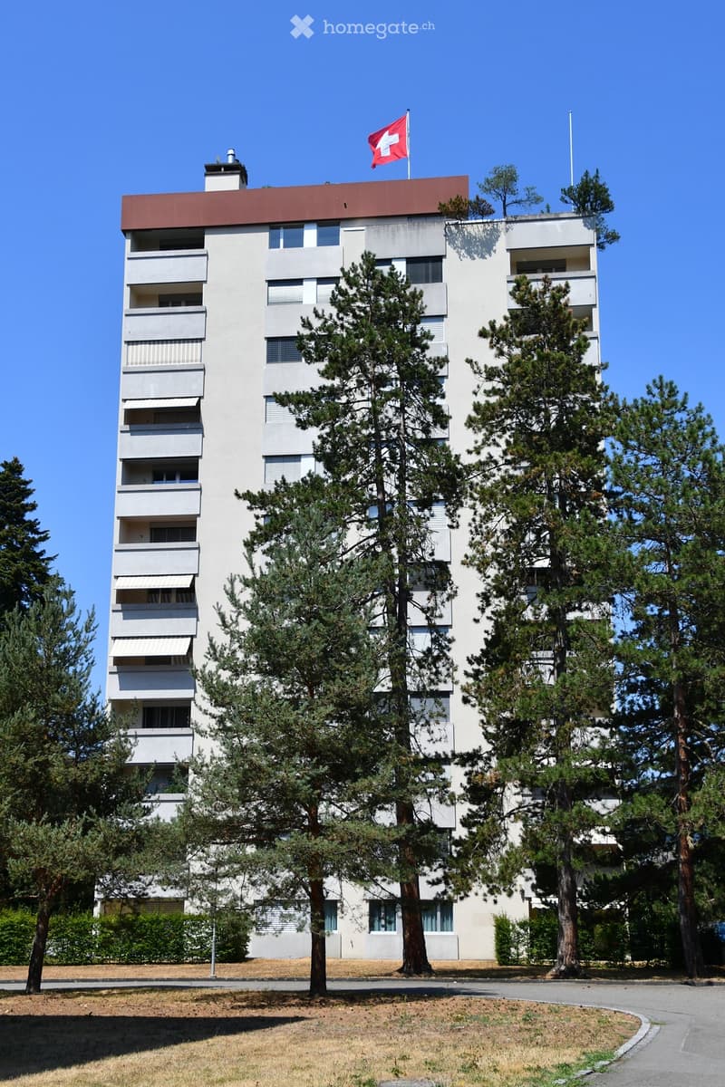 A modern, multi-story residential building with a white facade, balconies, and a red roof. The Swiss flag is displayed on top. Surrounded by tall pine trees and a paved driveway.