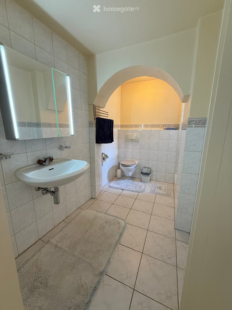 Modern bathroom with white tiles, a sink, toilet, mirror, and two bath mats.