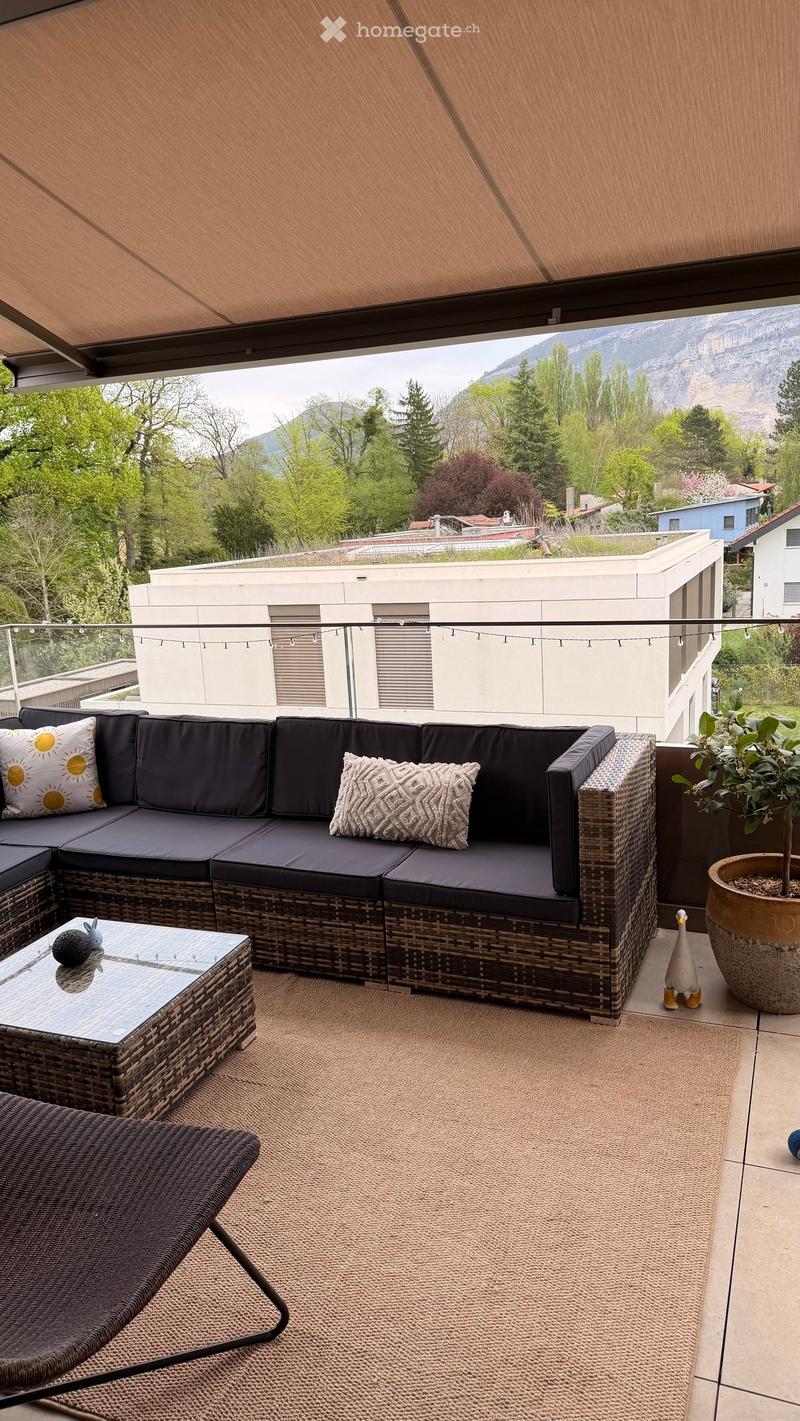 Balcony with a wicker sofa, coffee table, chair, rug, and potted plant, offering a scenic mountain view.