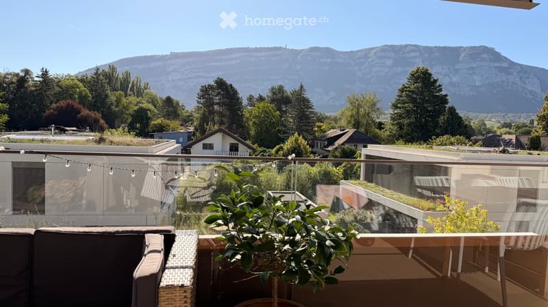 Balcony with a view of mountains, trees, and houses, featuring a wicker chair, a potted plant, and string lights.