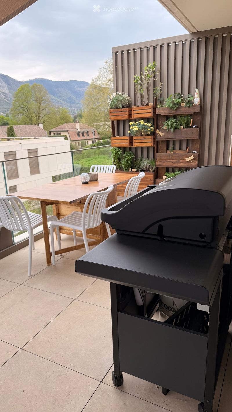 Balcony with a wooden table, white chairs, and a black grill, overlooking a scenic mountain view.