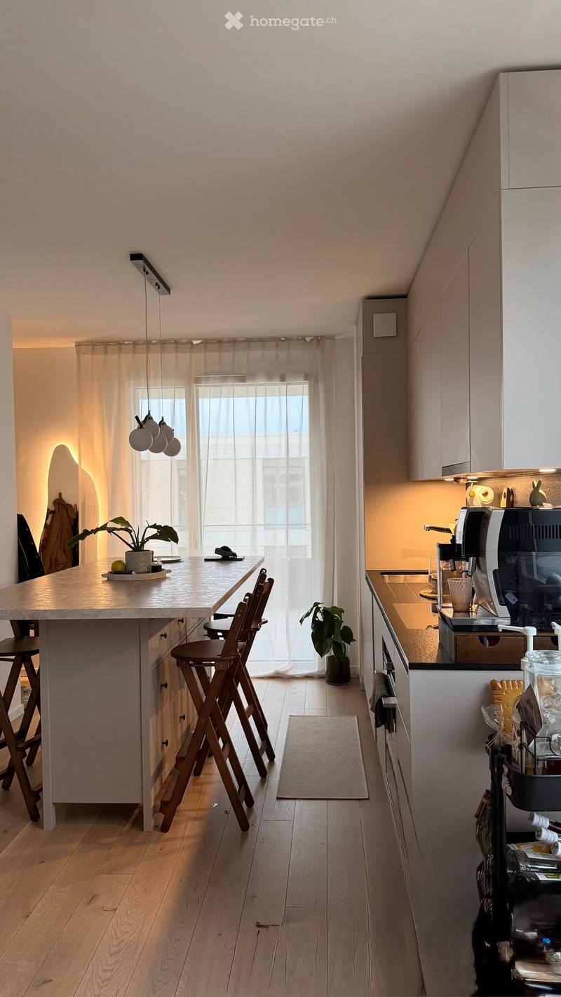 Modern kitchen with a central island, bar stools, coffee machine, and various appliances. Features wooden flooring, white cabinets, and large windows with sheer curtains.