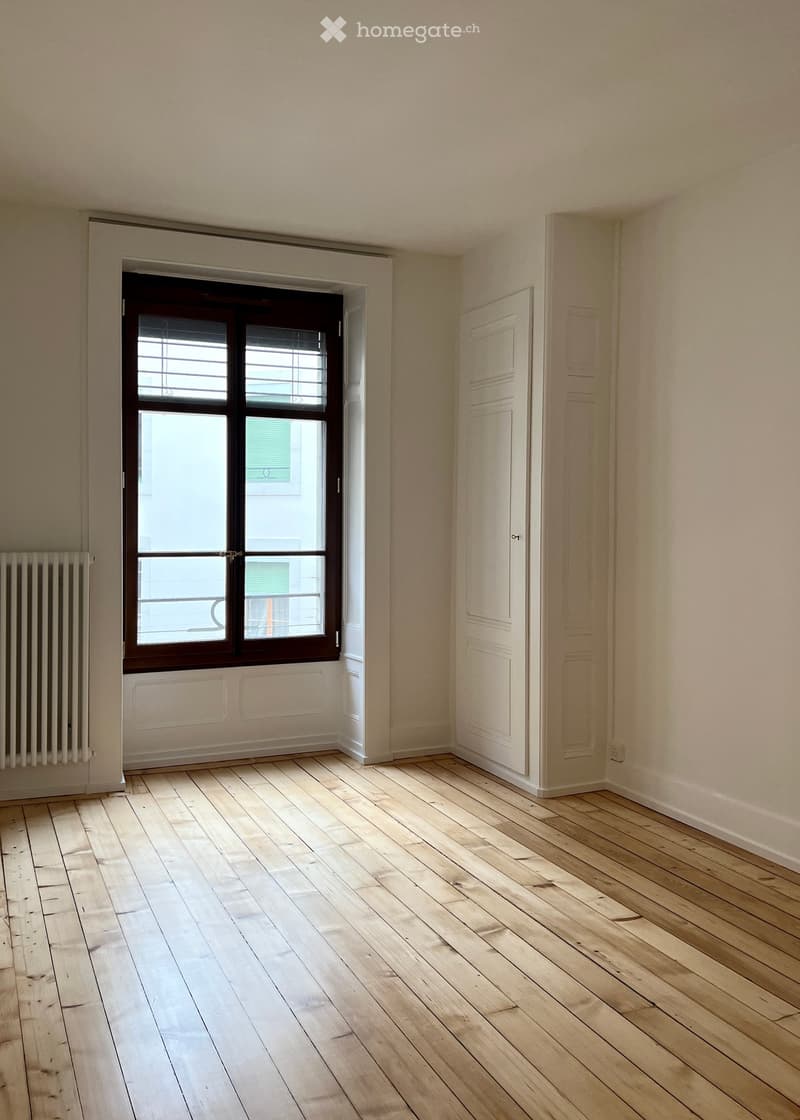 Empty room with wooden flooring, white walls, a window with brown frame, and a radiator on the left side.