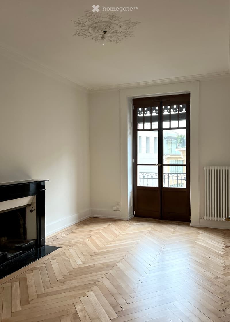 Empty room with light-colored walls, wooden herringbone flooring, a fireplace, and a large windowed door leading to a balcony.