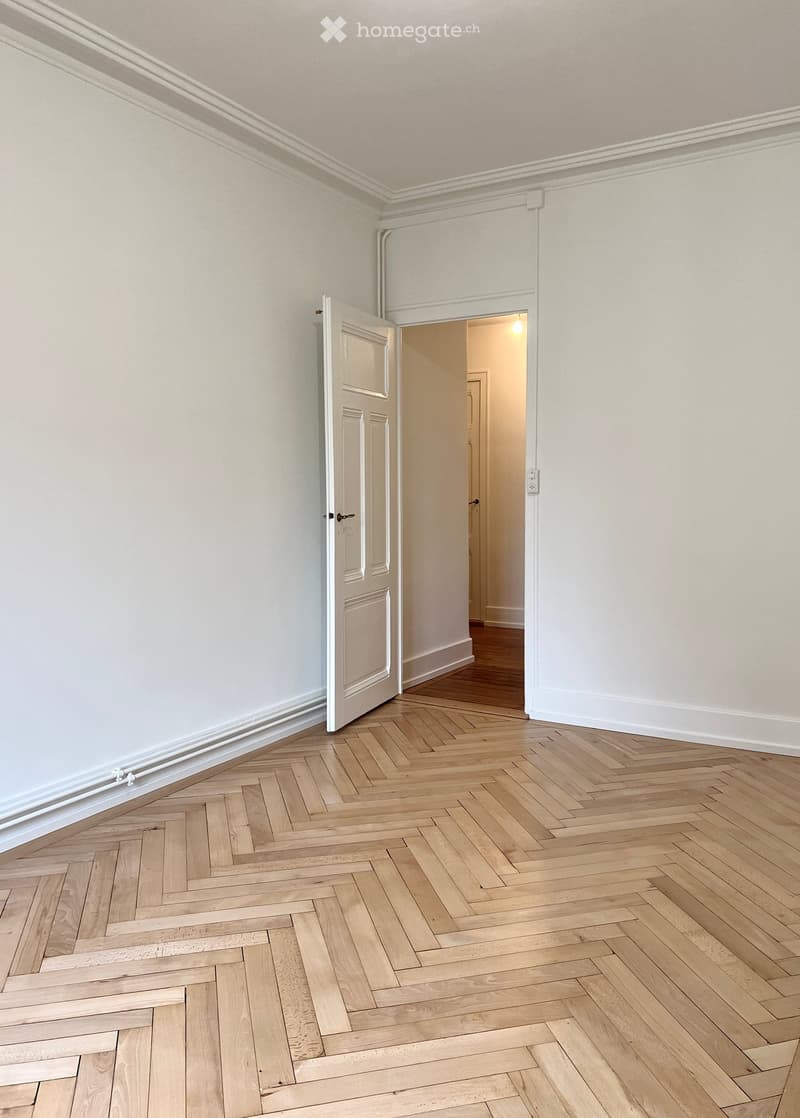 Empty room with light-colored walls, wooden herringbone flooring, and an open door leading to another room.