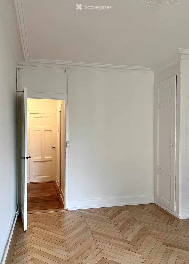 Empty hallway with wooden herringbone flooring, white walls, and a door leading to another room.