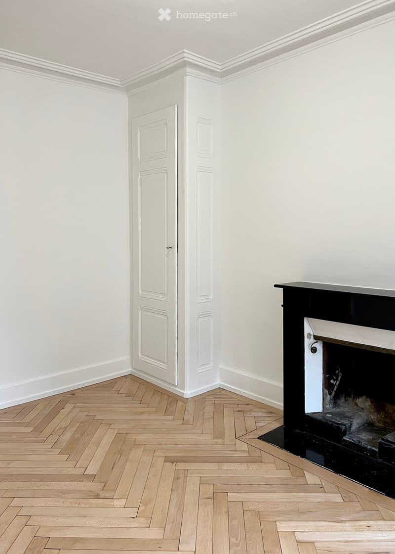 Empty room with white walls, wooden herringbone flooring, and a fireplace with a black mantel.